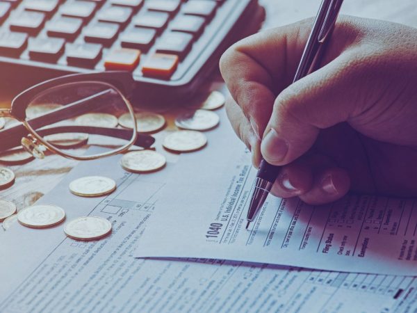 PESSOA ESCREVENDO EM PAPEL SOBRE A MESA COM UM ÓCULOS E ALGUMAS MOEADAS APOIADO