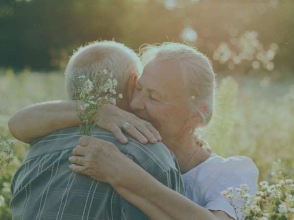 DOIS IDOSOS SE ABRANÇANDO EM UM GRAMADO